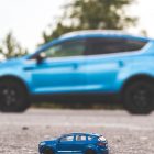 A close-up shot of a blue toy car on a road with a blurred blue SUV in the background, outdoors, during the day.