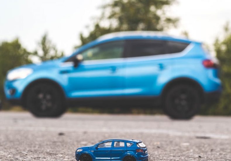 A close-up shot of a blue toy car on a road with a blurred blue SUV in the background, outdoors, during the day.
