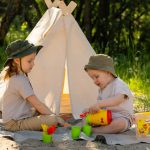 Two children enjoying summer playtime at a sandy campsite with a small tent.